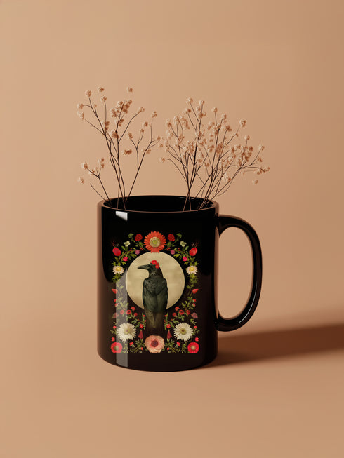A black ceramic mug featuring a gothic design of a raven against a golden full moon, framed by a floral wreath of coral-colored daisies, white chrysanthemums, and delicate botanical elements. The design includes rich green foliage and small red buds arranged in a symmetrical pattern. The mug has a classic C-shaped handle with a glossy finish.